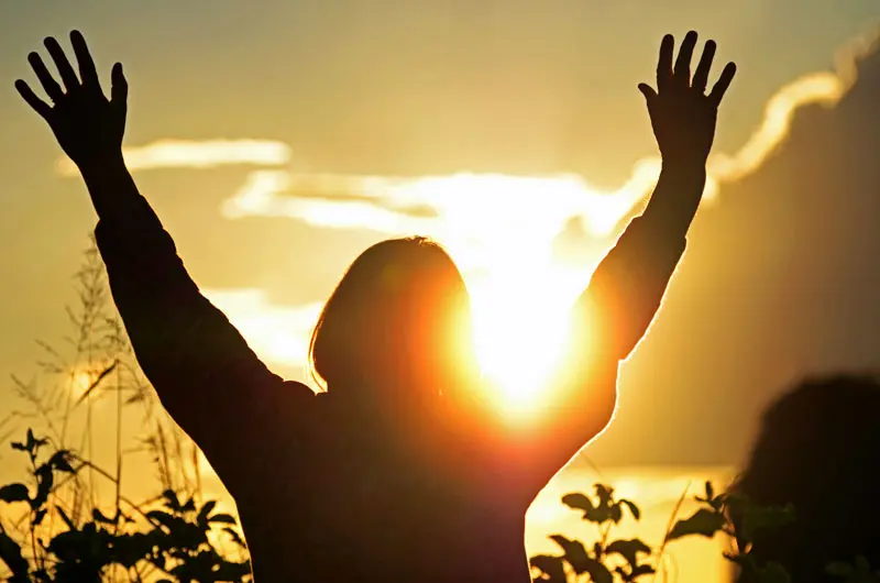 A woman raising her hands in the air with the sunrise behind her.