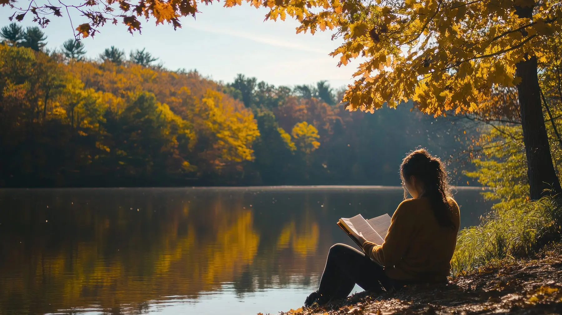 A woman reading a book by a lake in the fall season